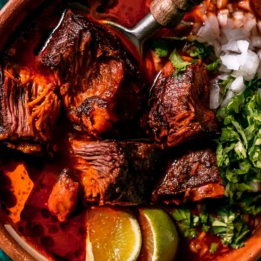A steaming bowl of authentic Mexican Birria served with fresh tortillas and toppings.